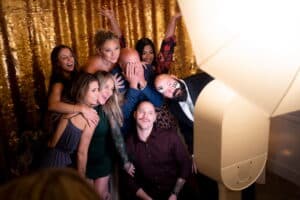 A group of seven people pose cheerfully in front of a glittery gold backdrop at an Orlando event, with some wearing formal attire. One person playfully hides their face while the others smile and gesture energetically towards the selfie station camera. A group of seven people pose cheerfully in front of a glittery gold backdrop at an Orlando event, with some wearing formal attire. One person playfully hides their face while the others smile and gesture energetically towards the selfie station camera.