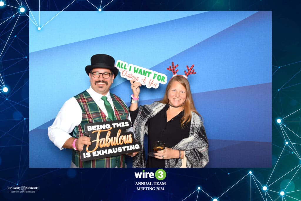 Two people strike a pose in front of a blue backdrop at the Wire 3 Annual Team Meeting 2024 using the digital photo booth. One sports a hat, glasses, and plaid vest with a Fabulous sign, while the other dons reindeer antlers and a shawl, holding an All I want for Christmas is You sign.