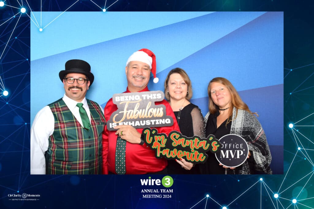 Four people pose in a digital photo booth, set against a vibrant blue backdrop. Theyre decked out in festive attire with playful props. A man wearing a Santa hat holds signs that declare, Being this fabulous is exhausting and Im Santas favorite, while another sign boasts Office MVP.