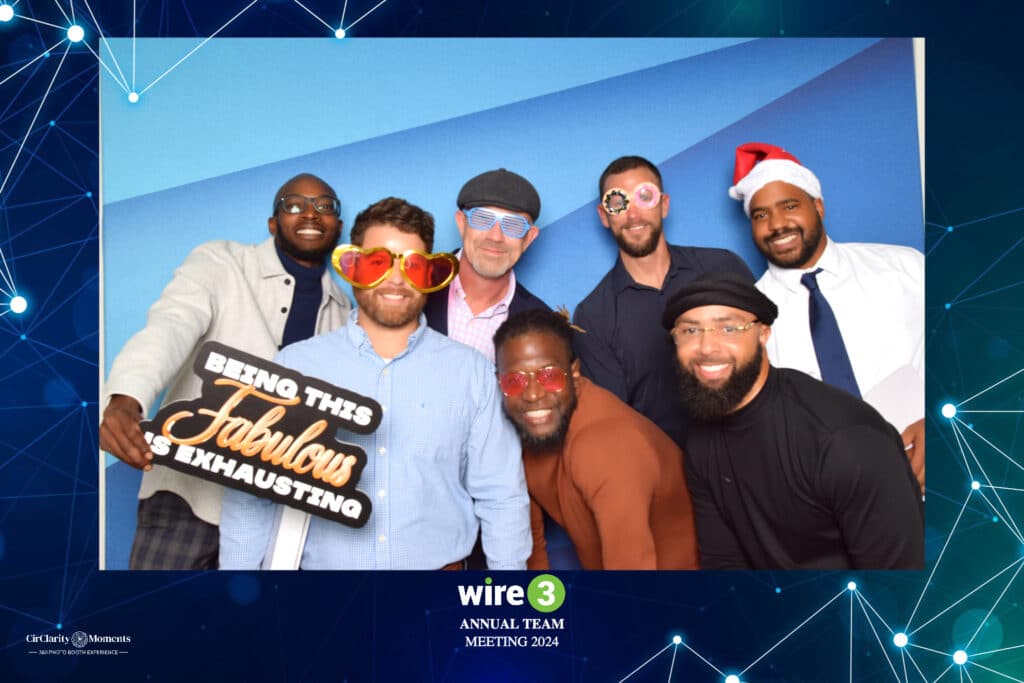 At the Wire 3 Annual Team Meeting 2024, a group of seven strikes playful poses in front of a digital photo booth. Theyre sporting oversized glasses and hats, with one donning a Santa hat. A sign reads, Being this fabulous is exhausting, capturing the spirited moment perfectly.