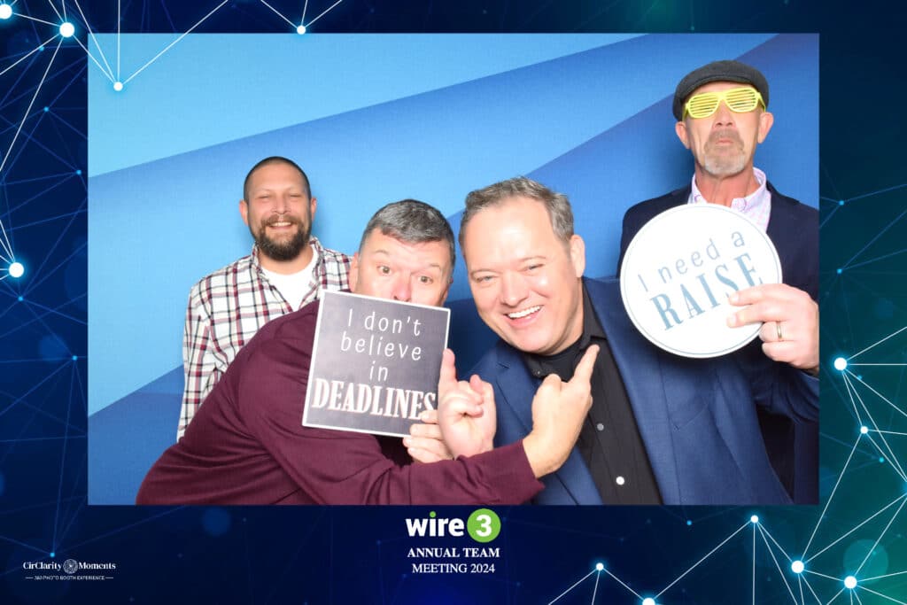 Four men pose playfully against a blue background at a digital photo booth. Two hold signs saying I dont believe in deadlines and I need a raise. One wears yellow striped glasses. Text at the bottom reads Wire 3 Annual Team Meeting 2024.