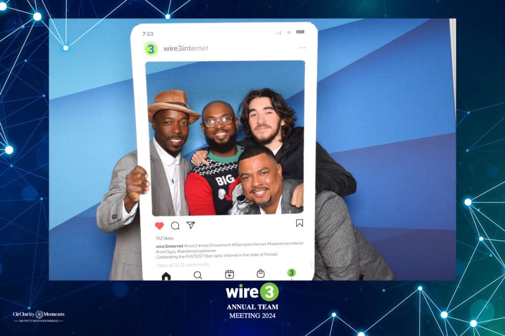 Four people smiling and posing with a large Instagram-style frame at the Wire 3 Annual Team Meeting 2024, giving off a digital photo booth vibe. Theyre set against a blue and green geometric backdrop, dressed in casual to semi-formal attire.