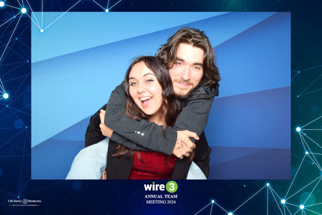 A smiling woman and man pose playfully in a digital photo booth, with the man hugging the woman from behind against a blue geometric backdrop. The image is framed with wire 3 Annual Team Meeting 2024.