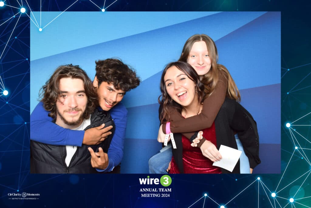 Four people are smiling and posing playfully against a blue backdrop at the Wire 3 Annual Team Meeting 2024. Two of them are piggybacking amidst a digital network design, capturing the fun moment in a lively Digital Photo Booth setting.