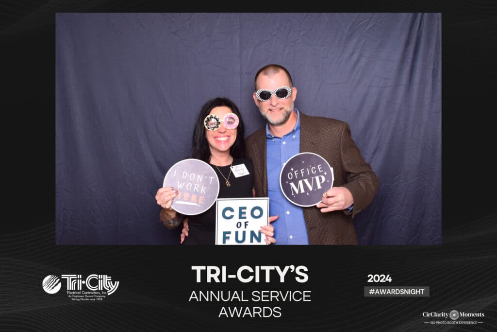 At Tri-Citys Annual Service Awards, two people beam while holding humorous signs—I dont work here and Office MVP—in front of a digital photo booth. Sporting novelty glasses, they capture the events fun spirit perfectly.