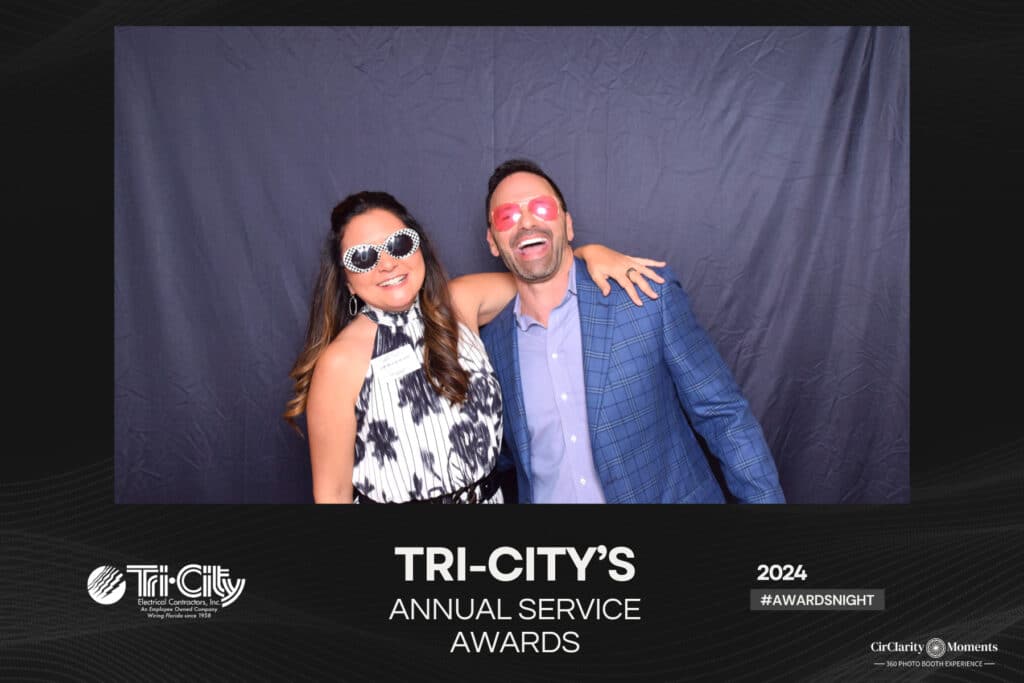 A woman and a man pose playfully in front of a dark backdrop at the Tri-Citys Annual Service Awards, captured perfectly by the digital photo booth. Both wear sunglasses; the woman in a patterned dress and the man in a blue suit, smiling with his arm around her shoulder.