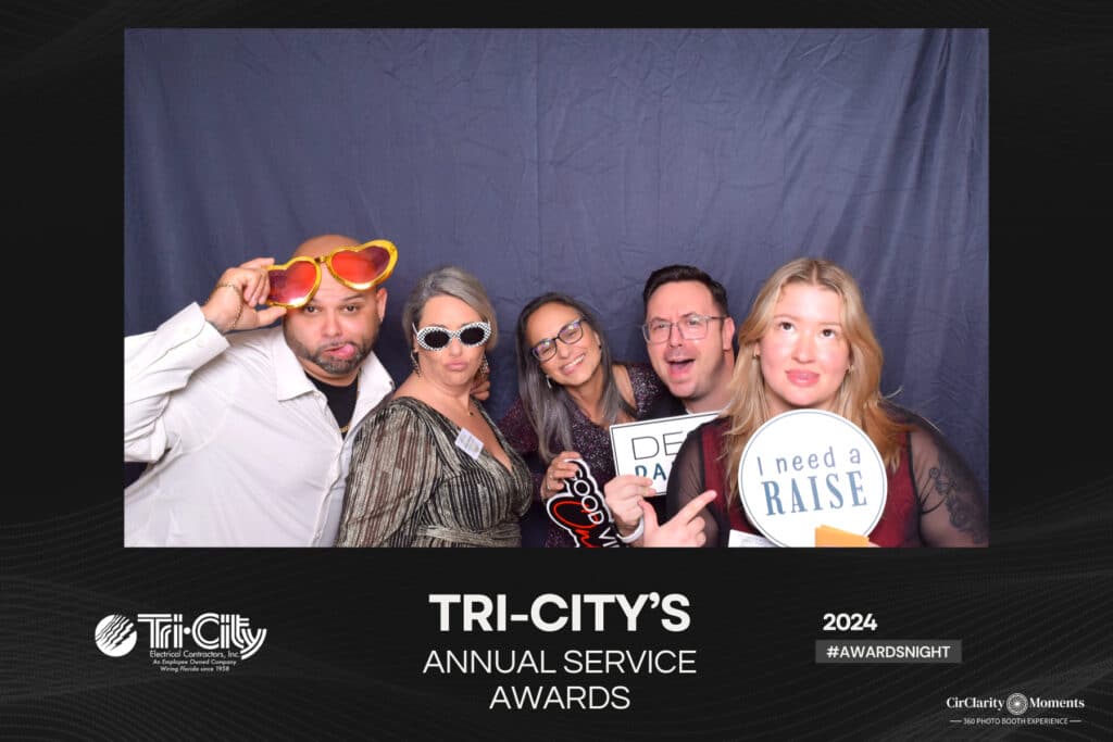 Five people pose playfully against a gray backdrop at Tri-City’s Annual Service Awards, captured perfectly in a digital photo booth. They hold quirky signs like I need a raise and wear fun accessories like sunglasses and heart-shaped glasses. Event details are at the bottom.