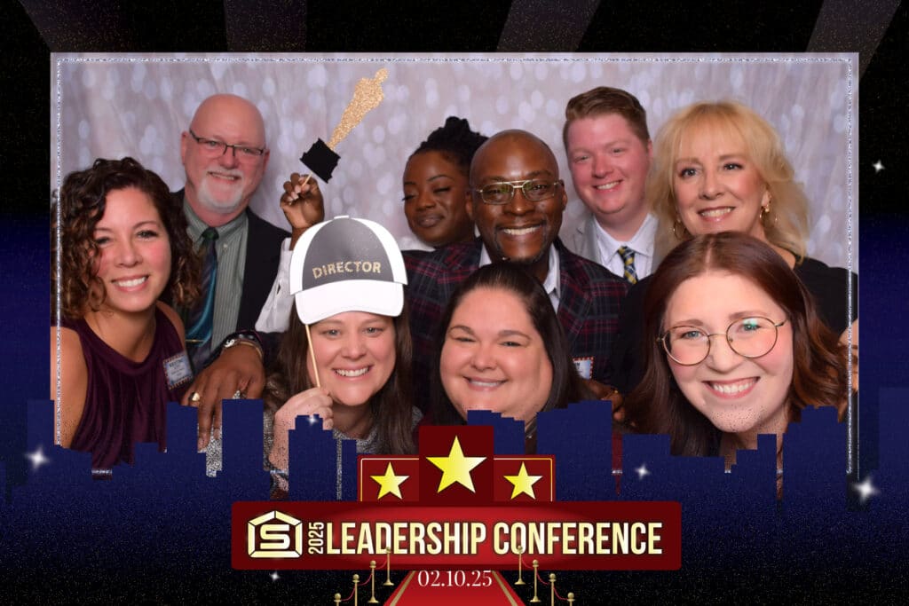 A group of nine smiling people at a leadership conference pose with fun props from a digital photo booth against a starry backdrop. Some hold a directors clapperboard and megaphone. Text reads 2025 Leadership Conference and 02.10.25 at the bottom.