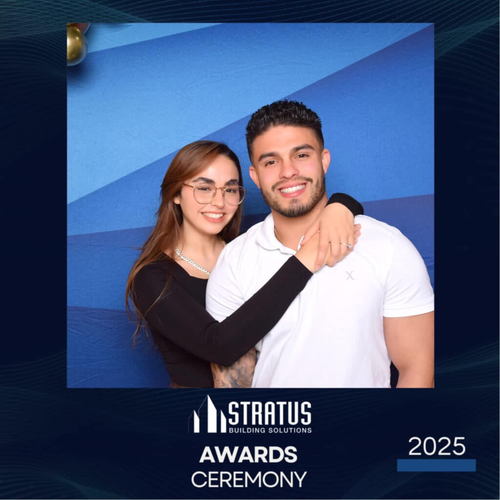 A woman and a man stand together smiling in front of a blue backdrop, captured perfectly by the digital photo booth. The woman has long hair and glasses, wearing a black top. The man has short hair, a beard, and is clad in a white polo shirt at the Stratus Building Solutions Awards Ceremony 2025.