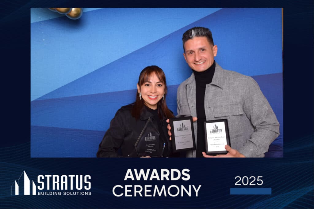 A man and woman smile while holding awards in front of a blue gradient backdrop at the 2025 Stratus Building Solutions Awards Ceremony. The event logo, name, and a digital photo booth add to the celebratory atmosphere.