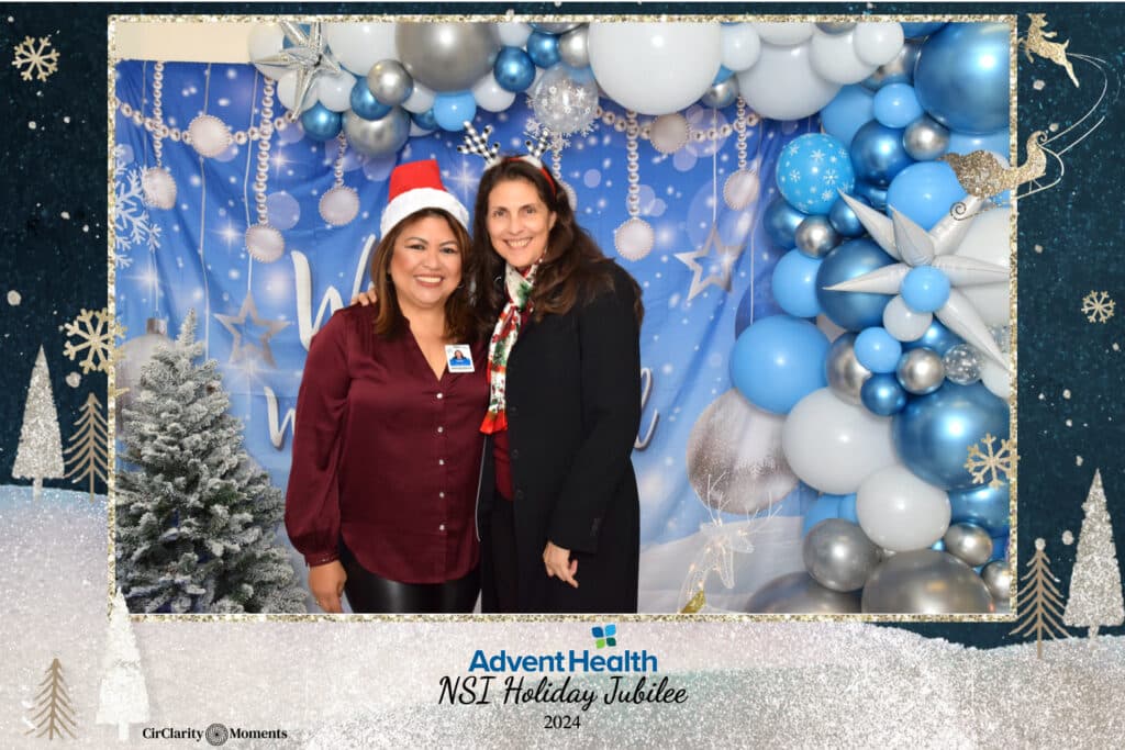 Two women beam at the AdventHealth NST Holiday Jubilee 2024, captured perfectly by the digital photo booth. They stand before a festive backdrop filled with blue and white balloons. One wears a Santa hat, the other a scarf, with snowflakes and trees adorning the frame.