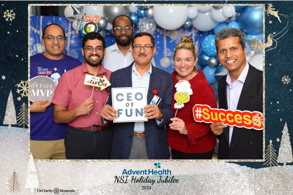 A group of six people smiling, posing in festive attire against a backdrop of balloons and a digital photo booth. They hold holiday-themed props, including signs that read CEO OF FUN, Success, and Mean One. Their joyful expressions convey a celebratory mood.