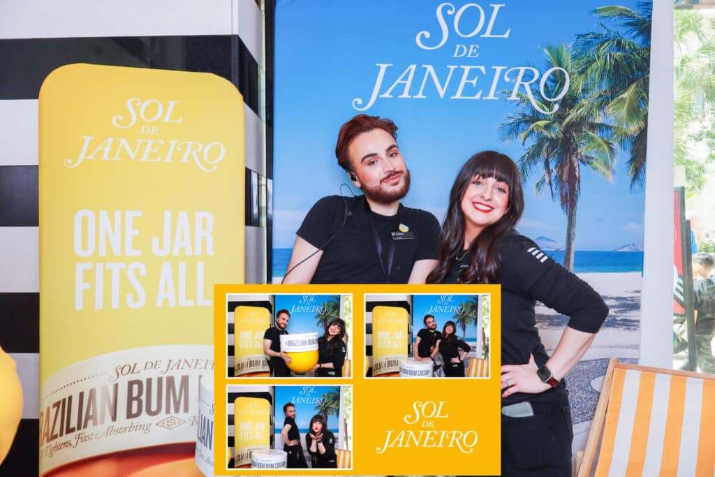 Two people smiling and posing at a Digital Photo Booth in front of a tropical Sol de Janeiro backdrop and product display, with palm trees, beach imagery, and a large yellow product stand that reads “One jar fits all.”.