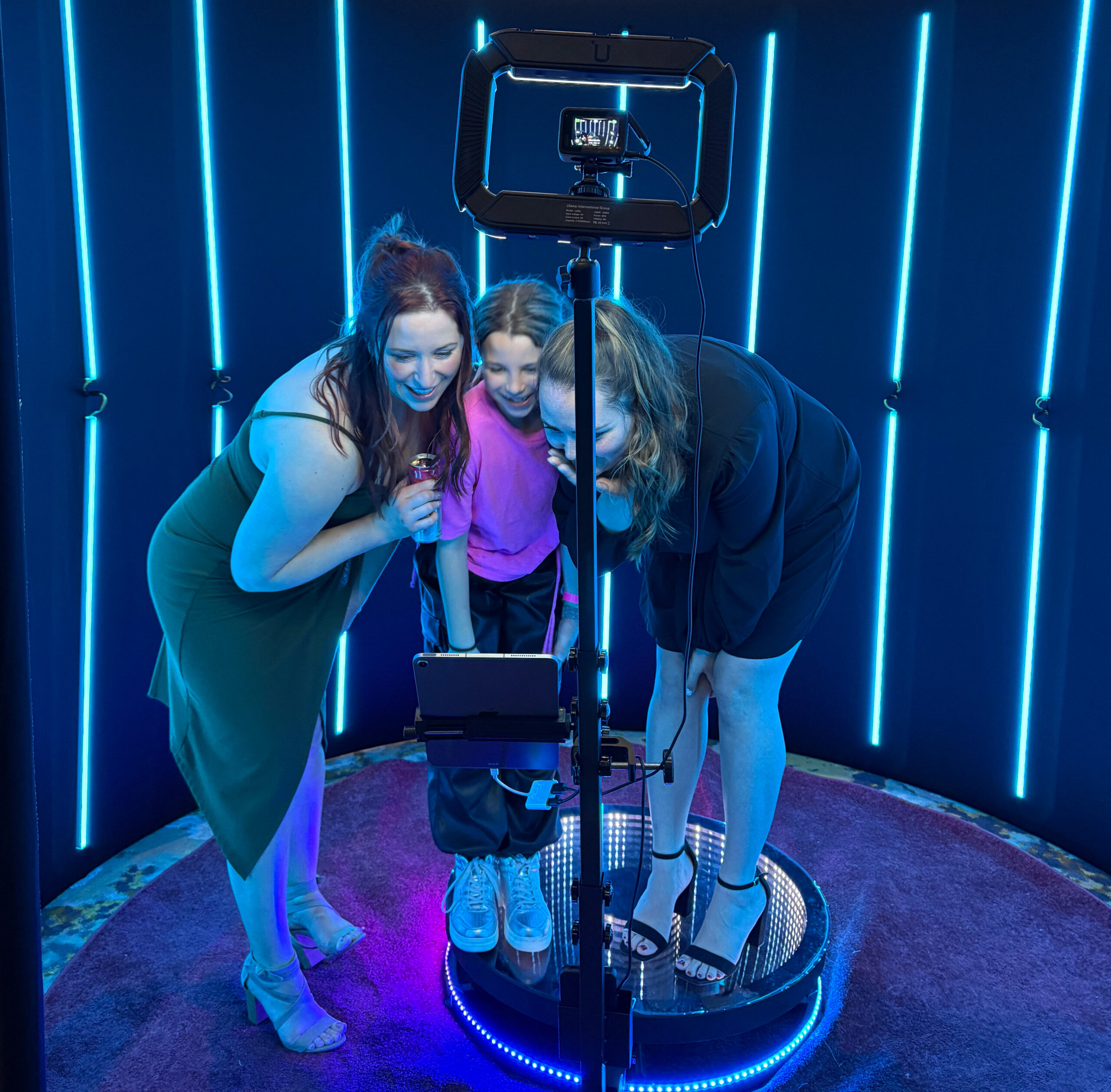 Three people stand on a circular platform inside a booth with neon blue lights. Theyre looking at a device on a stand. One holds a microphone, and they appear to be having fun. The background is dark, enhancing the blue glow.