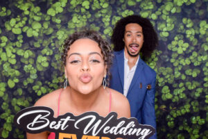 A woman with curly hair in a pink dress puckers her lips playfully, holding a sign that says Best Wedding Ever. A man with curly hair in a blue suit smiles in the background. The backdrop is decorated with a leafy pattern. A woman with curly hair in a pink dress puckers her lips playfully, holding a sign that says Best Wedding Ever. A man with curly hair in a blue suit smiles in the background. The backdrop is decorated with a leafy pattern.
