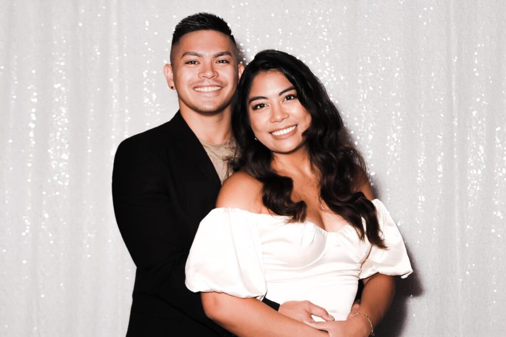 A smiling couple poses in front of a white, sparkly backdrop. The man, in a black jacket, stands behind the woman, who is wearing an off-the-shoulder white dress. They are both looking at the camera with cheerful expressions.