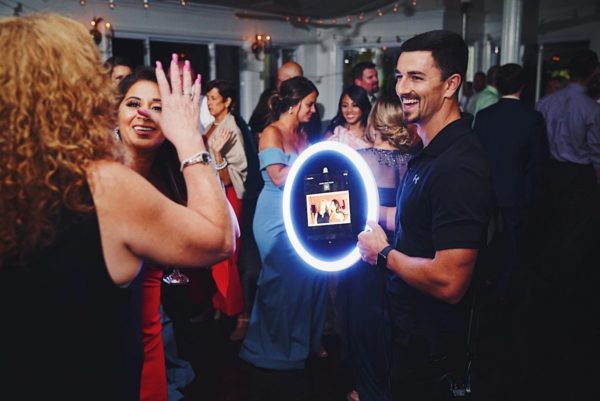 A man holds a circular light ring photo booth as smiling guests, dressed in formal attire, interact with it and enjoy themselves at a lively indoor party or event.