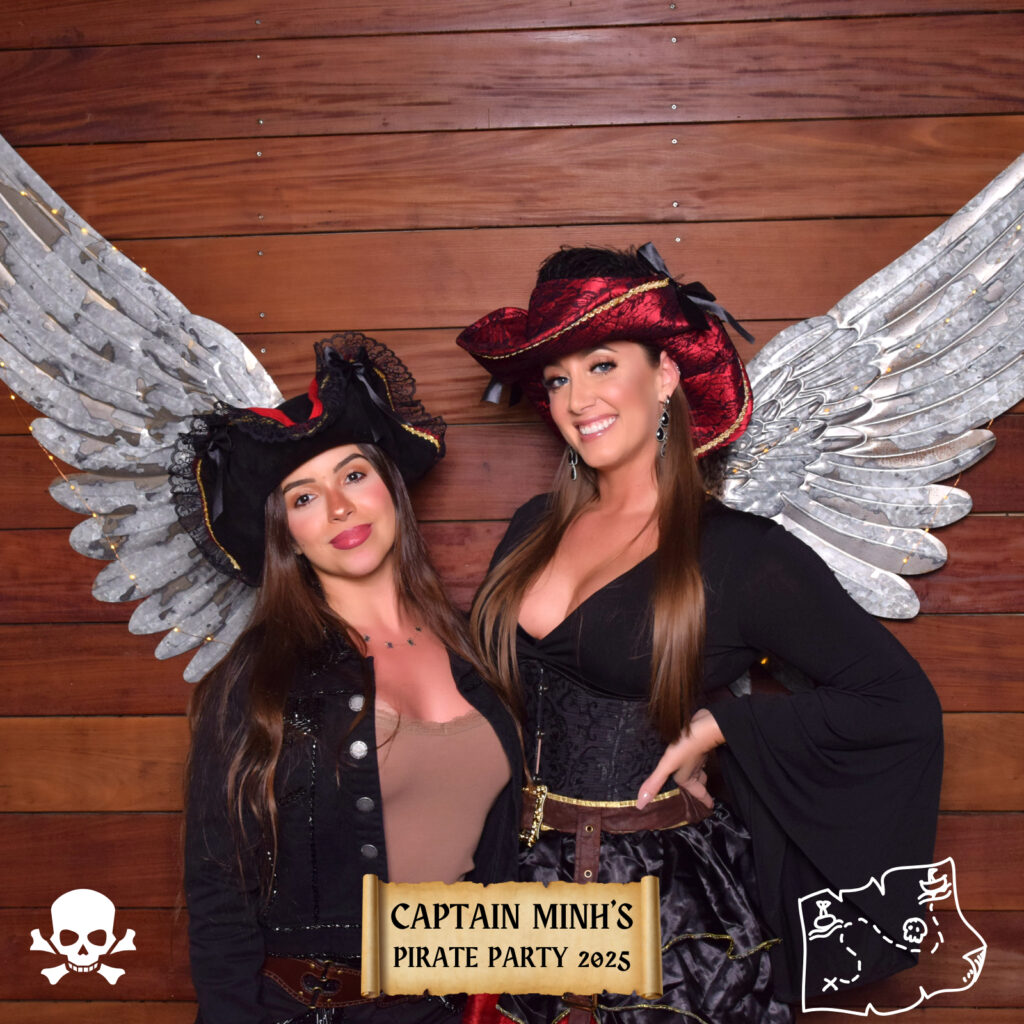Two women dressed as pirates pose in front of a wooden backdrop at a Digital Photo Booth. Both wear black hats, and the woman on the right has large silver wings. A banner reads Captain Minh&rsquo;s Pirate Party 2025, with skull and treasure map icons.