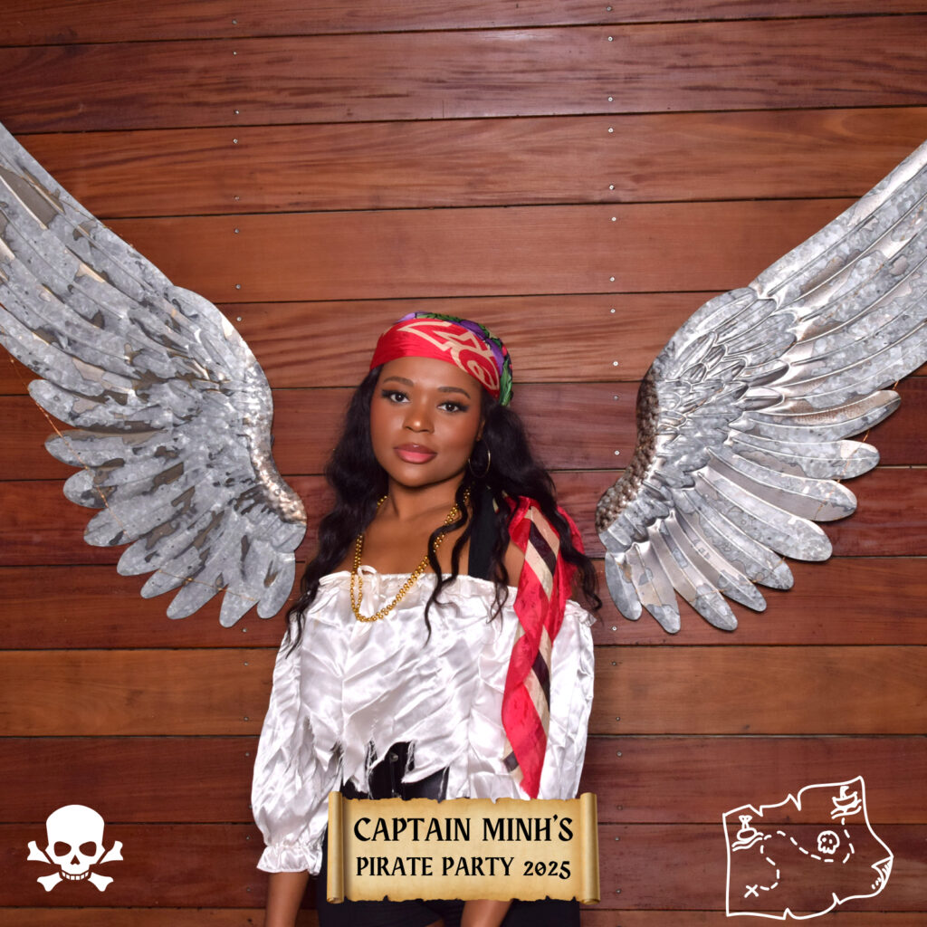 A woman in a pirate costume with metallic wings stands against a wooden wall beside a Digital Photo Booth. She wears a red headscarf, white blouse, and gold belt. Captain Minhs Pirate Party 2025 is written on a sign with pirate icons decorating the image.
