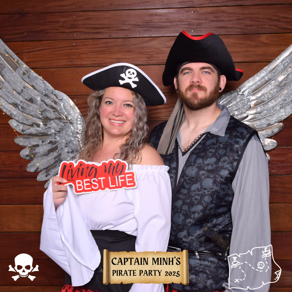 A woman and man dressed in pirate costumes stand in front of a wooden backdrop with metallic wings from a Digital Photo Booth. The woman holds a Living my BEST LIFE sign. The text reads Captain Minhs Pirate Party 2025 at the bottom.