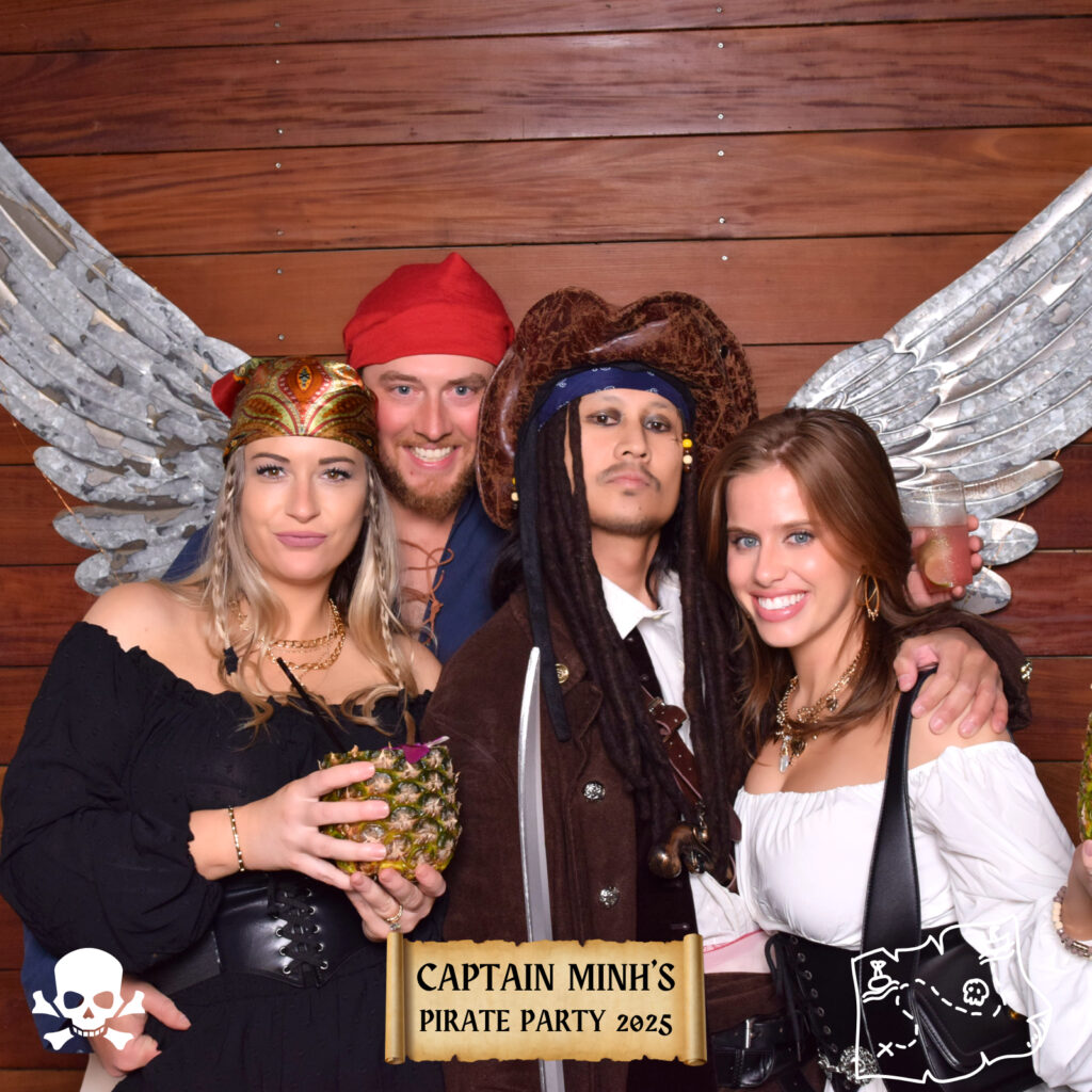 Four adults in pirate costumes pose together, smiling in front of a wooden wall with large silver wings at a Digital Photo Booth. A sign reads &ldquo;Captain Minh&rsquo;s Pirate Party 2025,&rdquo; and one person holds a drink in a pineapple cup.
