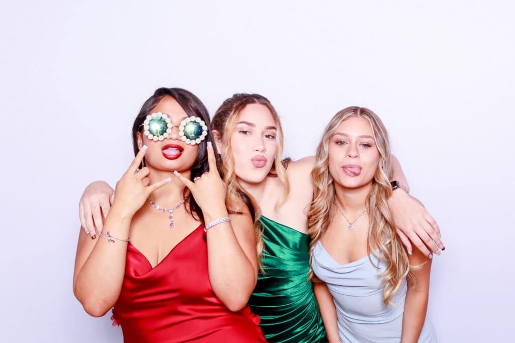 Three young women in formal dresses pose playfully against a white background, as if enjoying a Digital Photo Booth—one wears novelty glasses with green lenses while the others smile and stick out their tongues, arms wrapped around each other. Three young women in formal dresses pose playfully against a white background, as if enjoying a Digital Photo Booth—one wears novelty glasses with green lenses while the others smile and stick out their tongues, arms wrapped around each other.