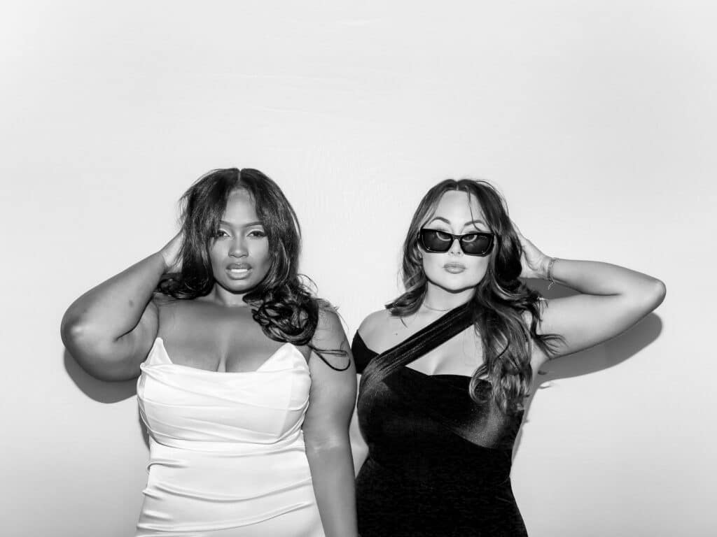 Two women pose confidently in front of a white background, captured by a Digital Photo Booth. One wears a light, off-shoulder dress, the other a dark, one-shoulder dress with sunglasses. Both have one hand touching their hair in this black and white shot. Two women pose confidently in front of a white background, captured by a Digital Photo Booth. One wears a light, off-shoulder dress, the other a dark, one-shoulder dress with sunglasses. Both have one hand touching their hair in this black and white shot.