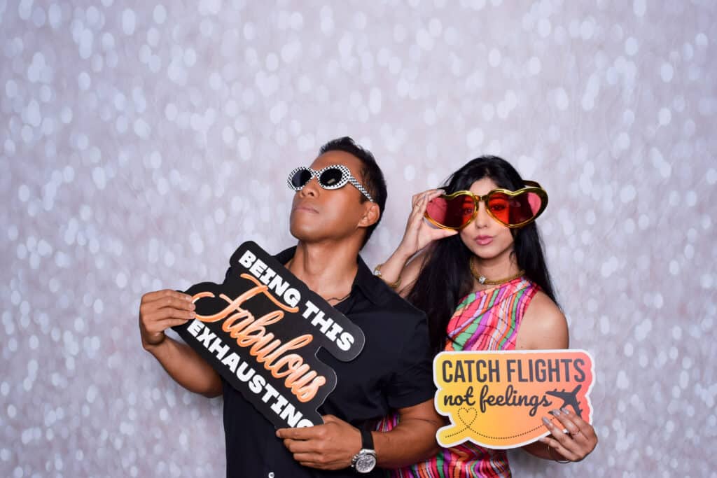 Two people pose playfully in front of a sparkly Digital Photo Booth backdrop. One holds a sign reading Being this fabulous is exhausting, the other holds a Catch flights not feelings sign—both rocking colorful, oversized sunglasses. Two people pose playfully in front of a sparkly Digital Photo Booth backdrop. One holds a sign reading Being this fabulous is exhausting, the other holds a Catch flights not feelings sign—both rocking colorful, oversized sunglasses.