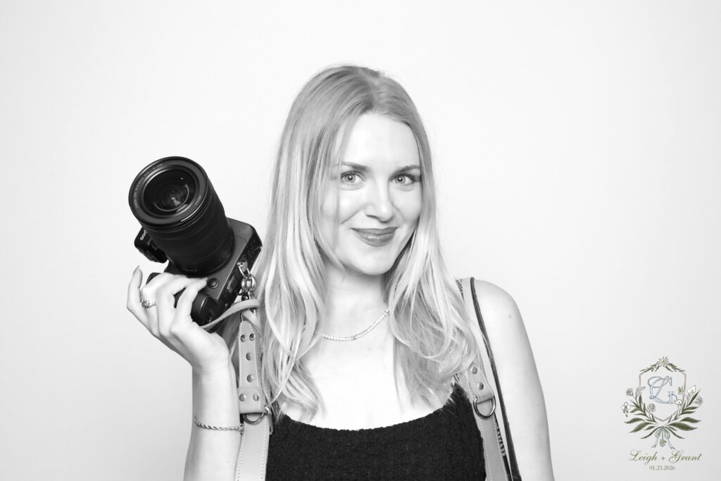 A smiling woman with long blonde hair holds a camera on her shoulder, posing against a plain light background. She wears a sleeveless top and necklace. A decorative logo and Digital Photo Booth branding appear in the bottom right corner.
