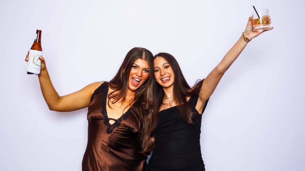 Two women smiling and posing in front of a white background, one holding a bottle and the other holding a glass with a straw, both with arms raised in celebration at a digital photo booth.