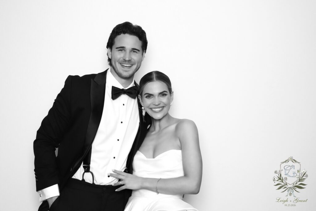 A smiling couple in formal attire poses against a plain background, captured by a Digital Photo Booth. The man wears a tuxedo with a bow tie, and the woman wears a strapless dress. A floral logo with Leigh &amp; Forrest appears in the bottom right corner.