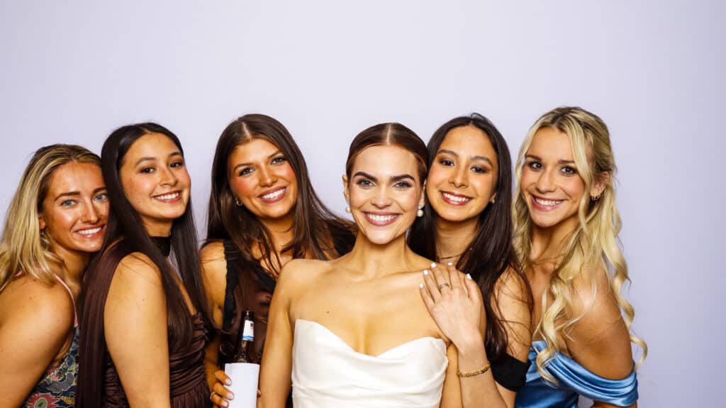 Six women pose closely together, smiling at the camera in front of a plain, light-colored background. The woman in the center wears a white strapless dress, likely the bride, as they enjoy a fun moment captured by a Digital Photo Booth.