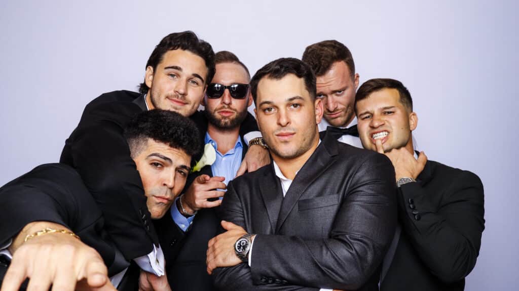 Six men in suits pose together against a plain light background, some smiling or making playful expressions. One crosses his arms while another points at the camera, capturing the fun atmosphere of a formal event in a Digital Photo Booth.