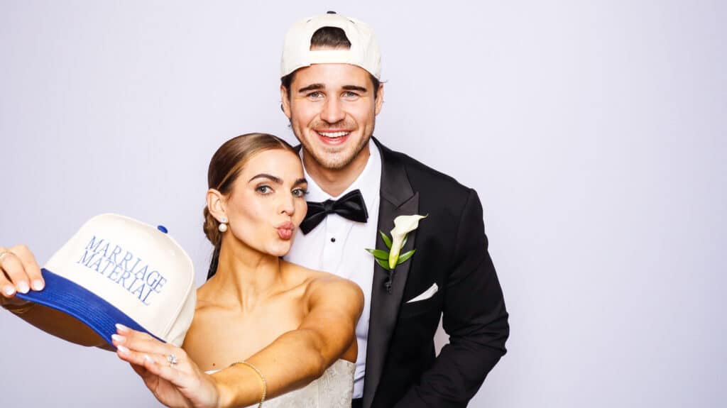 A bride and groom pose together in a Digital Photo Booth, both wearing white caps backward. The bride holds out a cap that says MARRIAGE MATERIAL and makes a playful kissy face, while the groom smiles in his tuxedo with a white boutonniere.
