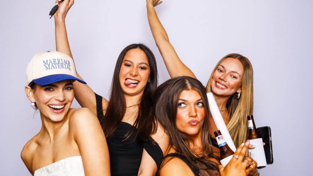 Four young women pose playfully in front of a light backdrop, smiling and making silly faces at a digital photo booth. One wears a Marriage Material hat, and two hold beer bottles, enjoying a fun celebration or party moment together.