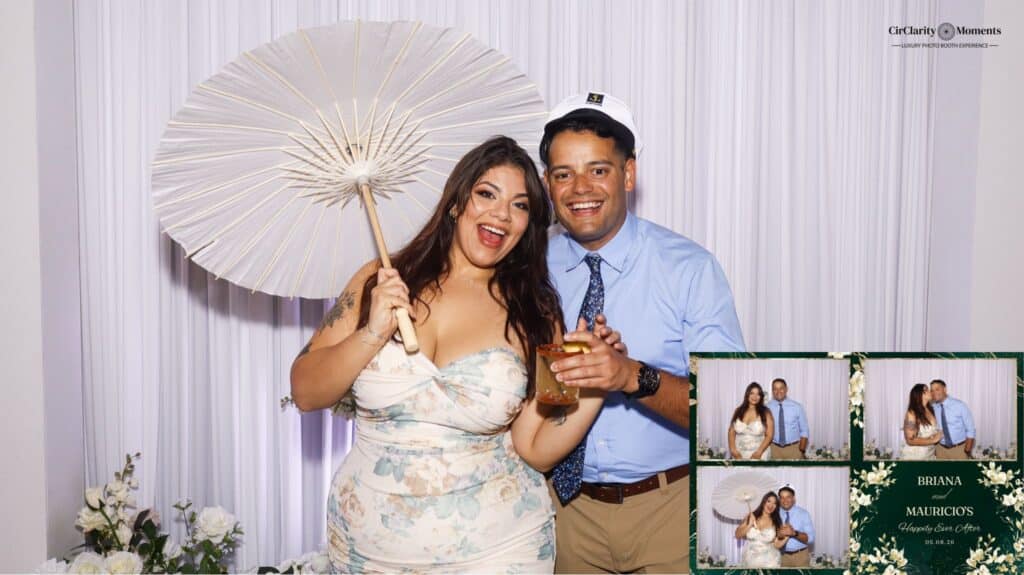 A smiling woman in a floral dress holds a white parasol and a drink, standing next to a man in a blue shirt and hat. They pose happily at the Digital Photo Booth, with flowers and a collage of smaller photos on the white backdrop.