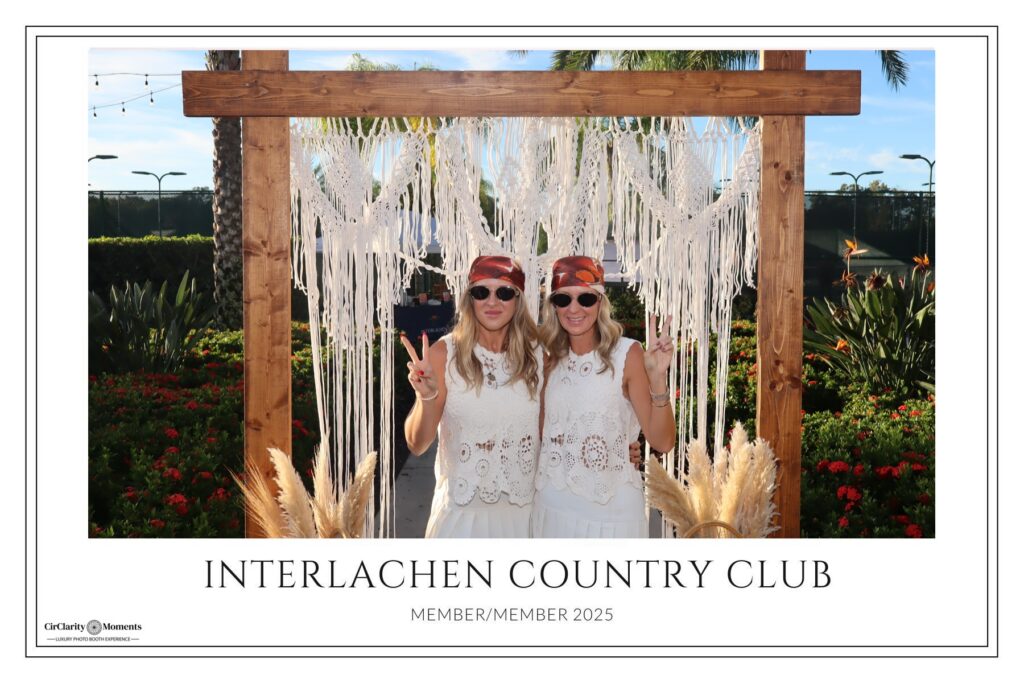 Two women in white lace outfits and headbands pose under a wooden arch with macram&eacute; decor, making peace signs. Outdoors with palm trees and flowers, they enjoy the moment at Interlachen Country Club&rsquo;s Digital Photo Booth.