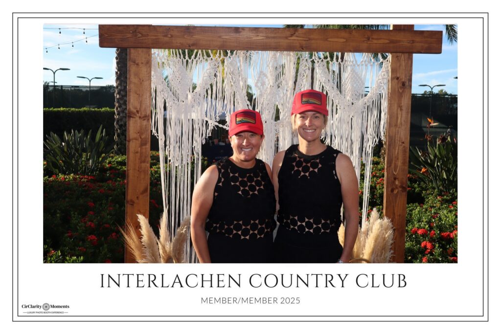 Two women in matching black dresses and red hats smile together in front of a wooden frame decorated with macram&eacute;, plants, and tall grasses at Interlachen Country Club&rsquo;s Digital Photo Booth. Text reads Member/Member 2025.