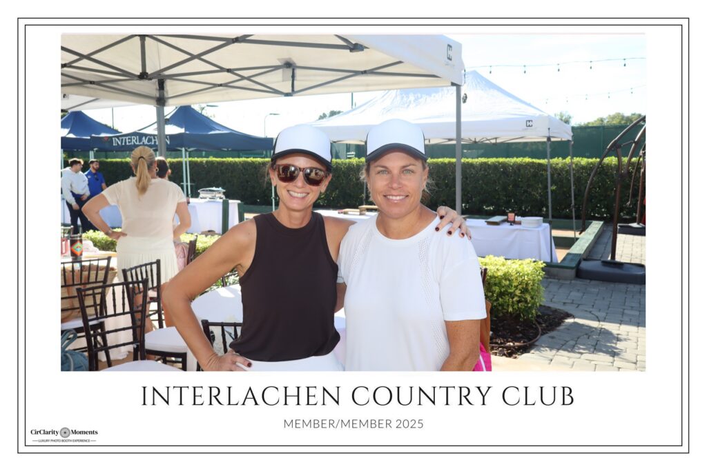Two women in white and black outfits and white caps pose together, smiling under a tent by the Digital Photo Booth at an outdoor event. A sign reads Interlachen Country Club with Member/Member 2025 below.
