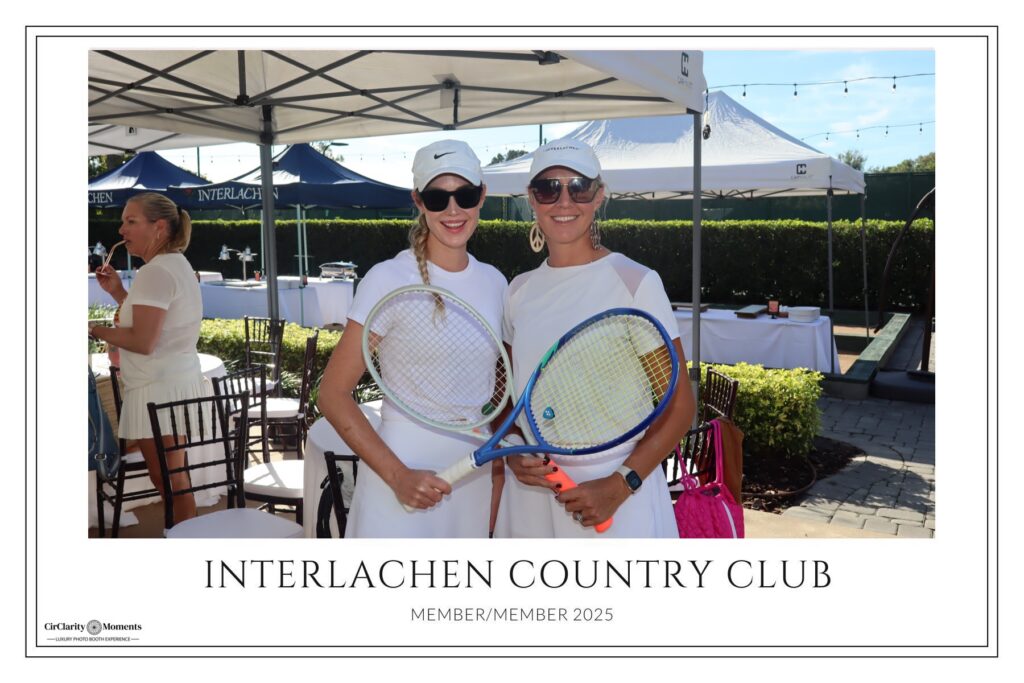 Two women in white tennis outfits and visors smile while holding tennis racquets at an outdoor event under tents, with a Digital Photo Booth nearby. The background features tables, chairs, and greenery. Text reads: Interlachen Country Club Member/Member 2025.