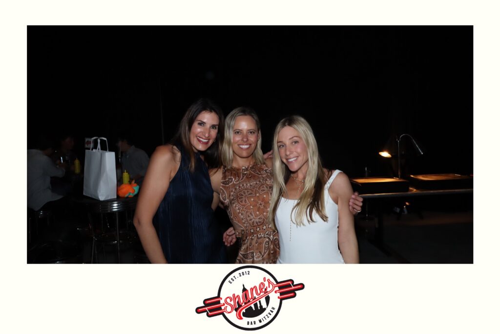Three women smiling and posing together at an indoor event, captured by a Digital Photo Booth. Two men sit in the background on the left. The image has a white border and Shanes Bar Mitzvah logo at the bottom.