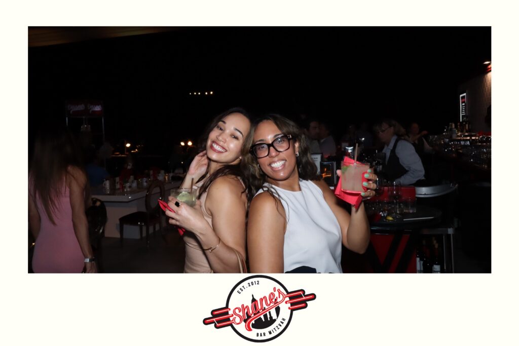 Two women smiling and posing with cocktails at a lively bar event, captured by a Digital Photo Booth. One woman makes a playful face while both hold drinks; people and a bar are visible in the background. The Shaker logo is at the bottom.