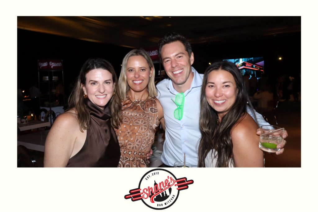 Four people stand close together, smiling at the camera at an event. One person holds a drink. The background features tables, lights, a Digital Photo Booth, and a Shane's Bar Mitzvah logo at the bottom.