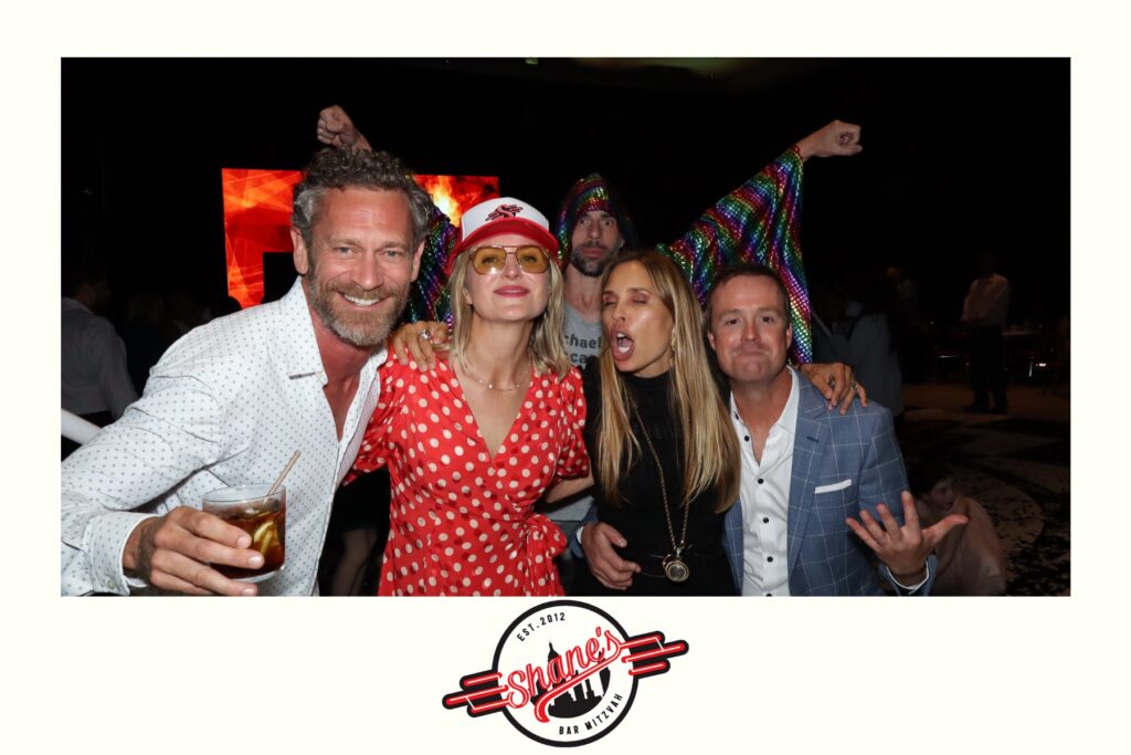 Five adults pose together at an event. Four stand in the foreground, smiling and making playful expressions, while one person in the back, using a Digital Photo Booth prop, wears a colorful shiny outfit with arms raised. The Shanes Bar Mitzvah logo is at the bottom.